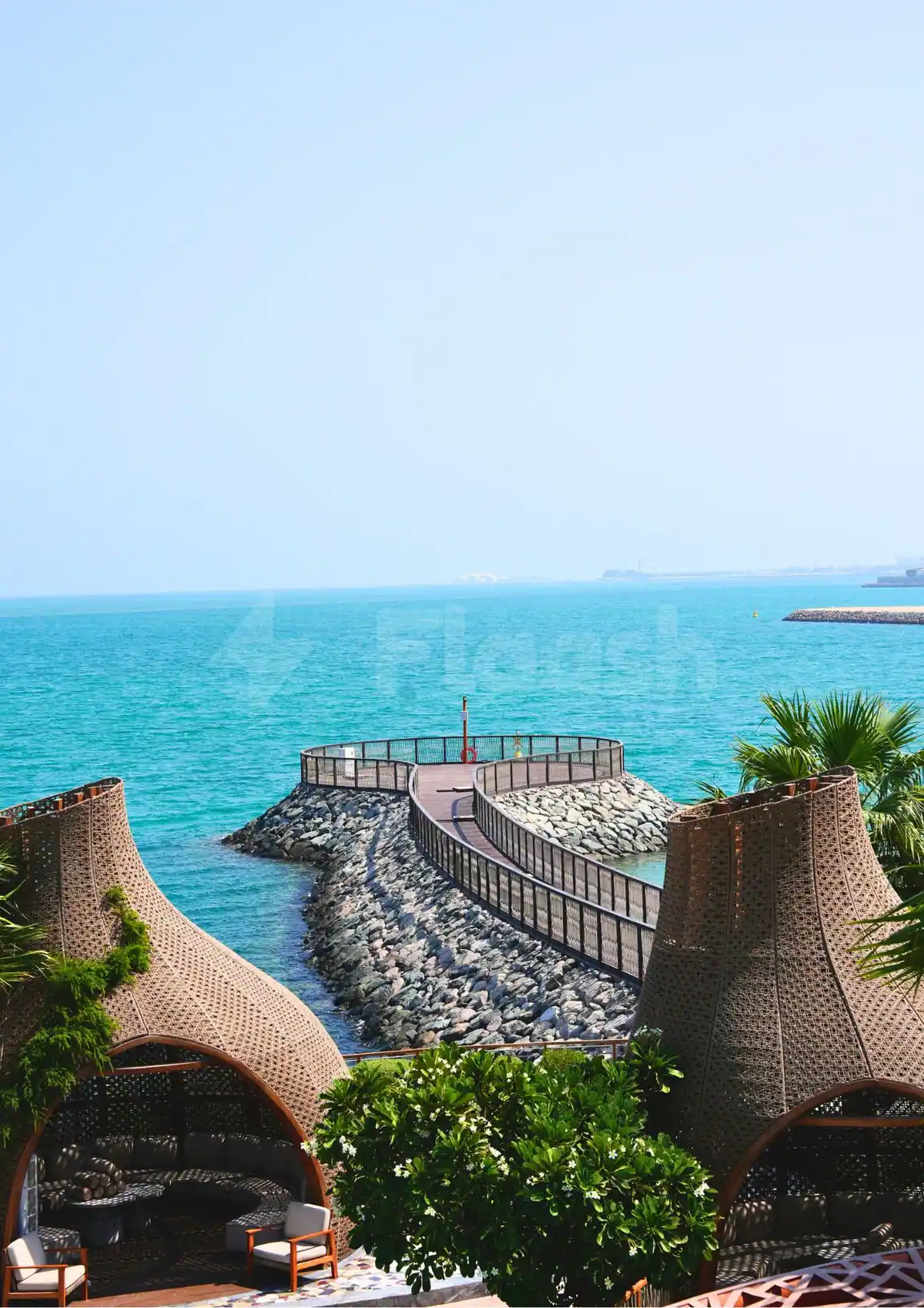 Terrace at Sushisamba Lusail featuring woven wicker cabanas and a curved wooden walkway over a rocky breakwater