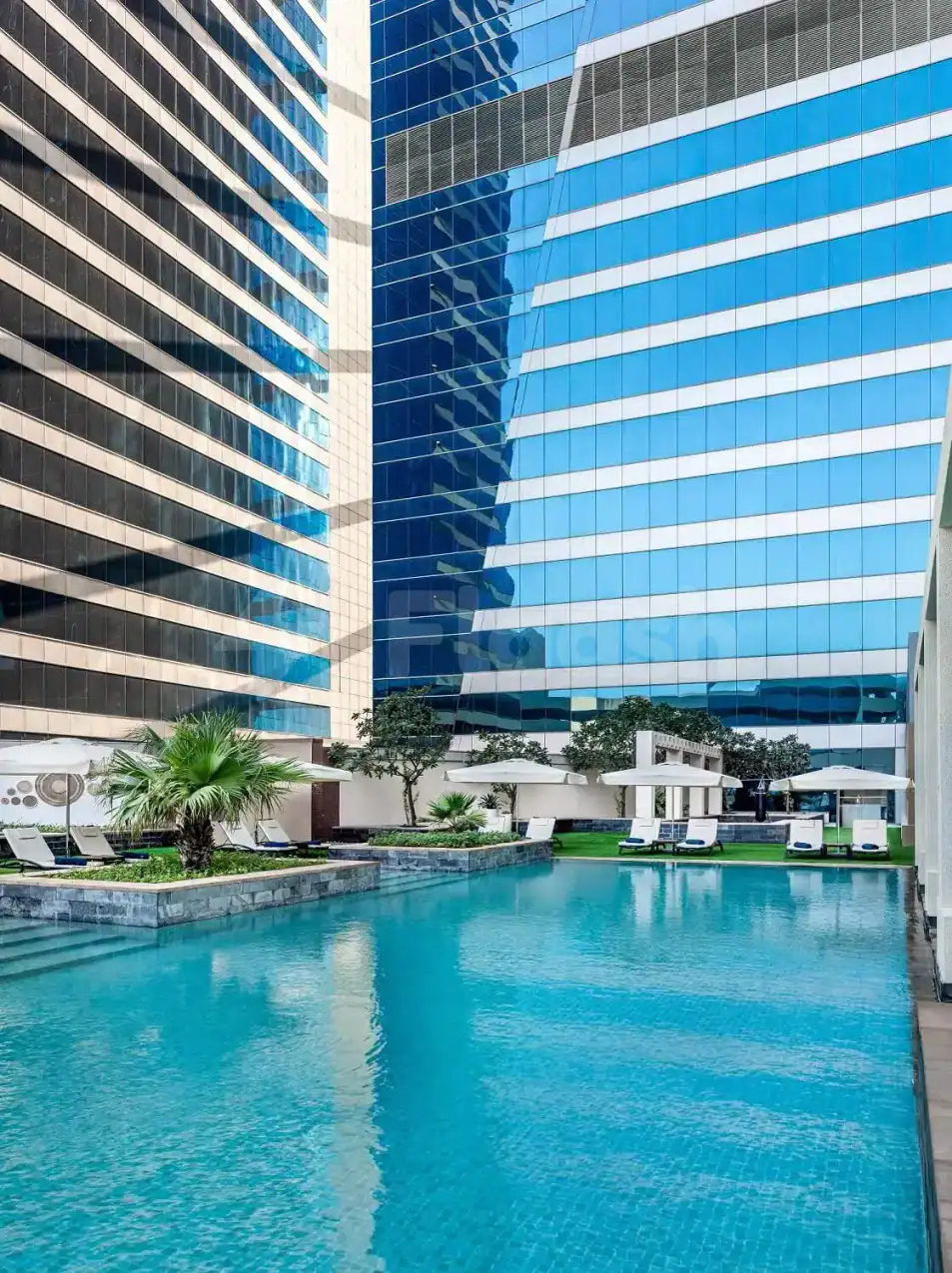 Rectangular swimming pool at The H Hotel Dubai with turquoise water, white lounge chairs, and glass-facade buildings nearby