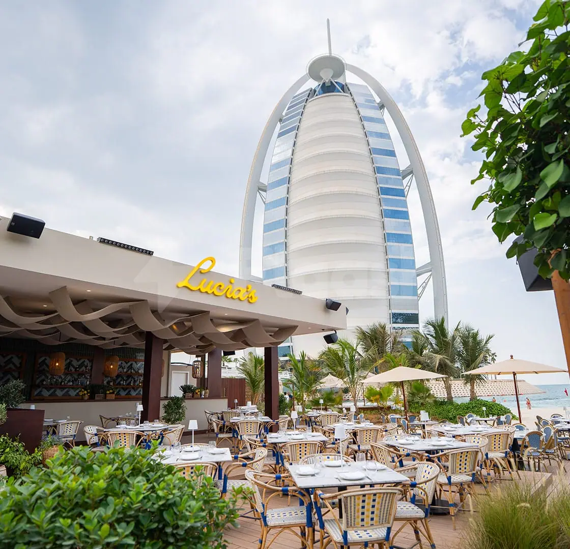 Outdoor dining area at Lucia's Mare Restaurant Dubai with wicker chairs, white tables, and the Burj Al Arab