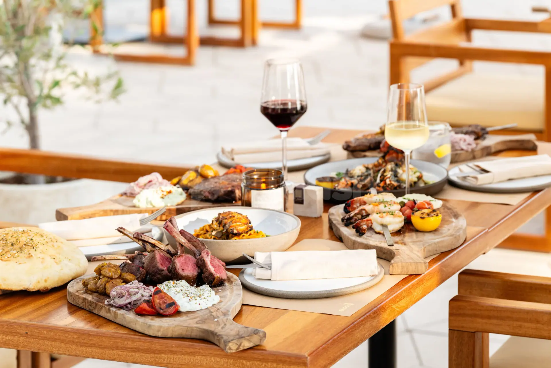 Wooden dining table at Ergon Deli & Cafe Abu Dhabi set with grilled lamb chops, shrimp, and wine glasses