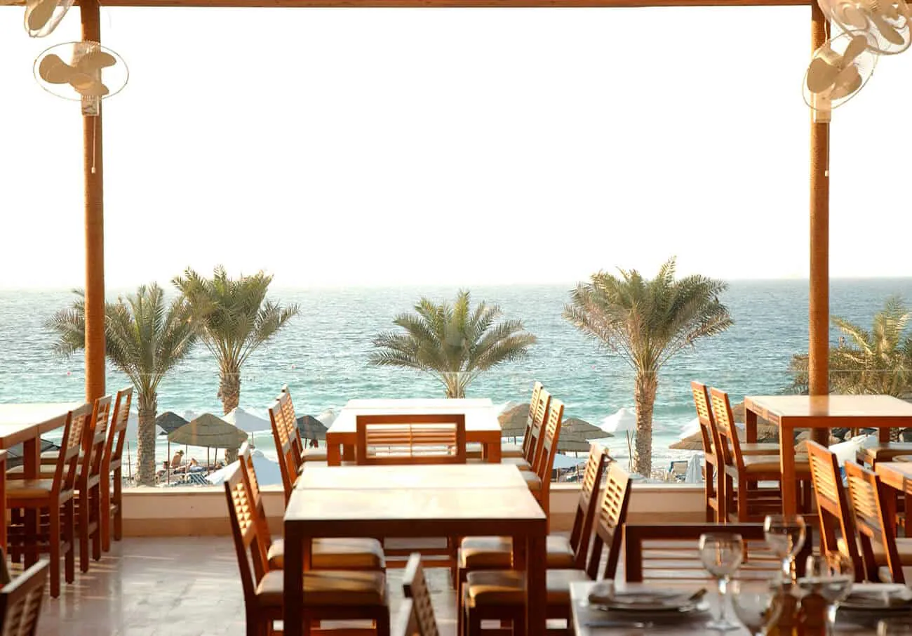 Open-air dining area at Flooka Restaurant Dubai with wooden tables and chairs, palm trees, and a direct ocean