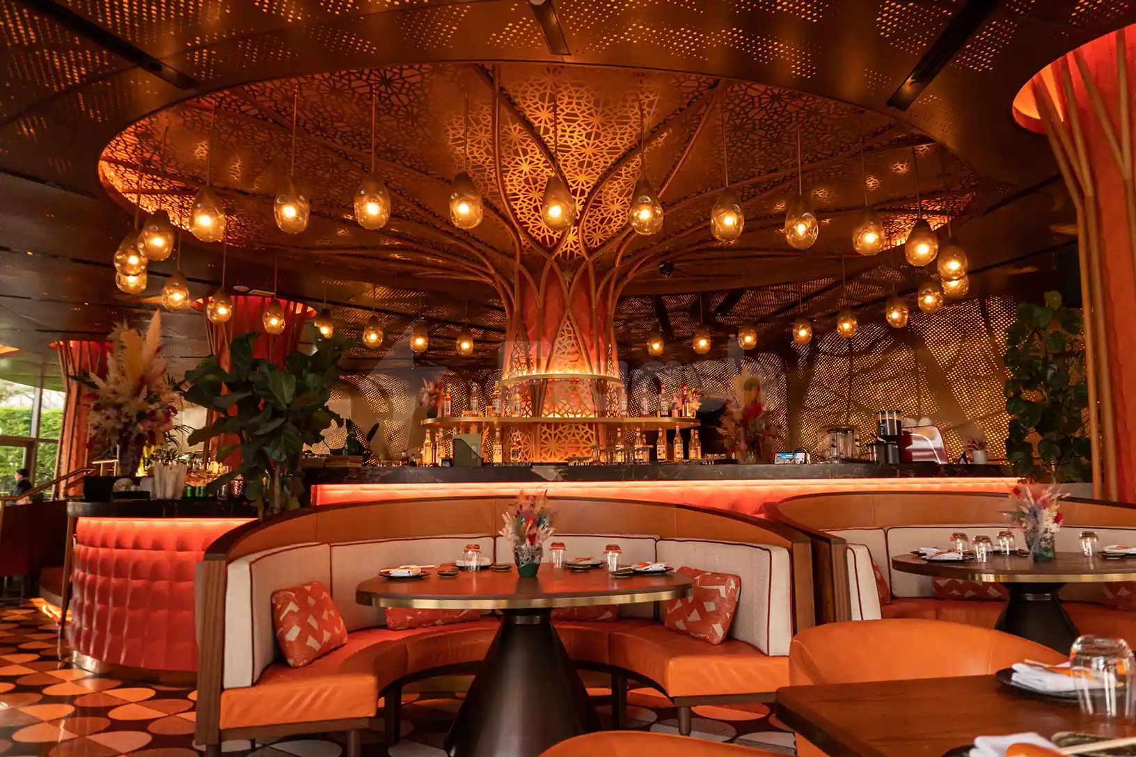 Restaurant interior at Sushisamba Lusail featuring a central ornate laser-cut metal canopy and curved booth seating with orange