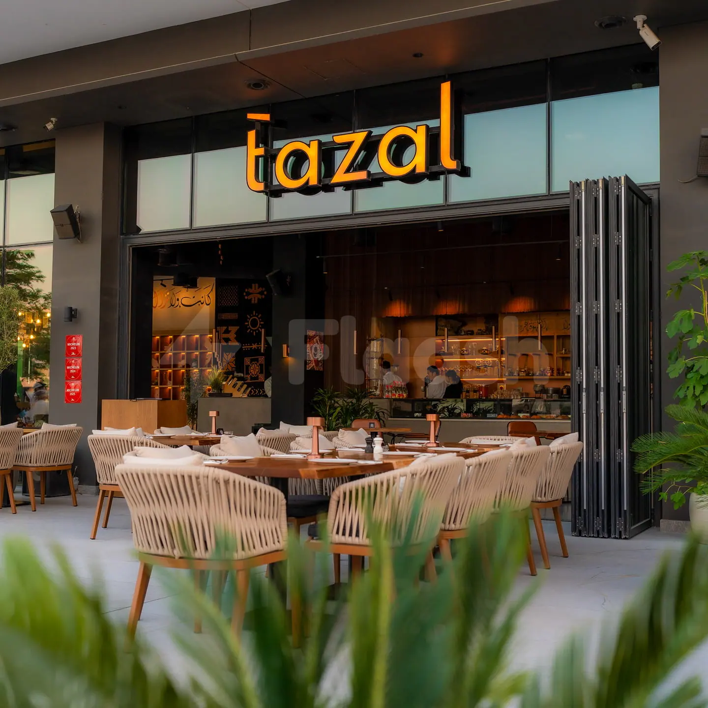 Outdoor dining area at Tazal Restaurant Abu Dhabi featuring woven chairs, wooden tables, and an open glass facade