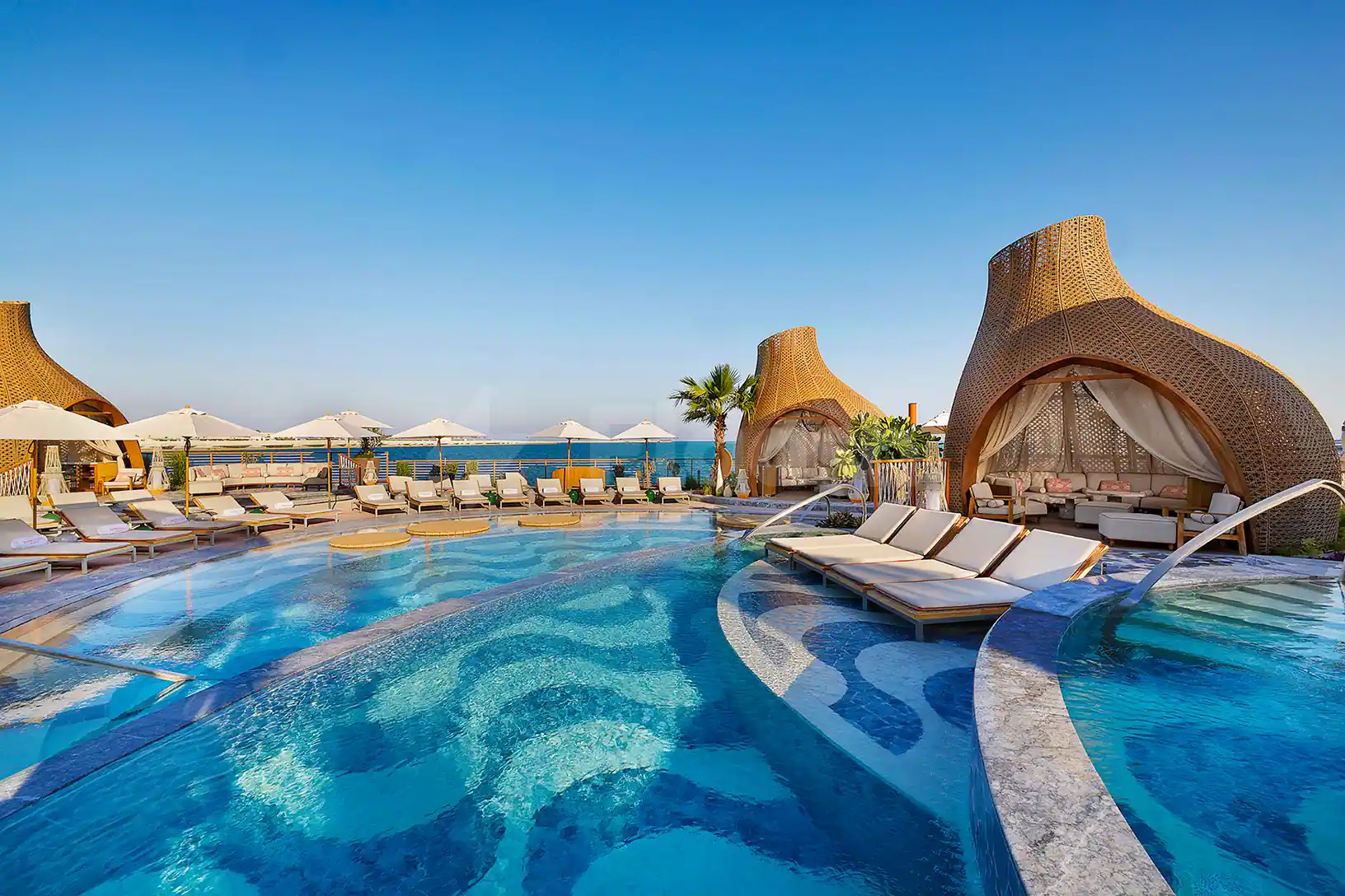 Curved swimming pool at Sushisamba Lusail in Lusail, featuring mosaic tile flooring and woven wicker cabanas with sheer