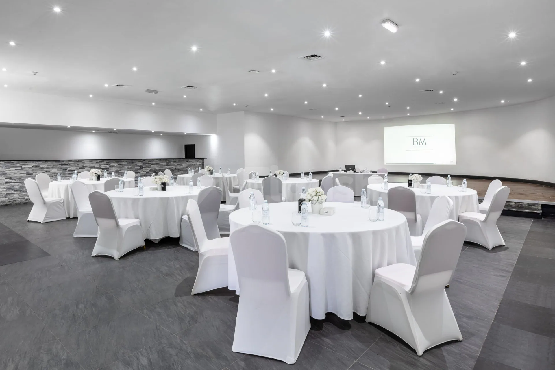 Ballroom at BM Beach Resort Ras Al Khaimah with round tables, white chair covers, and a projector screen