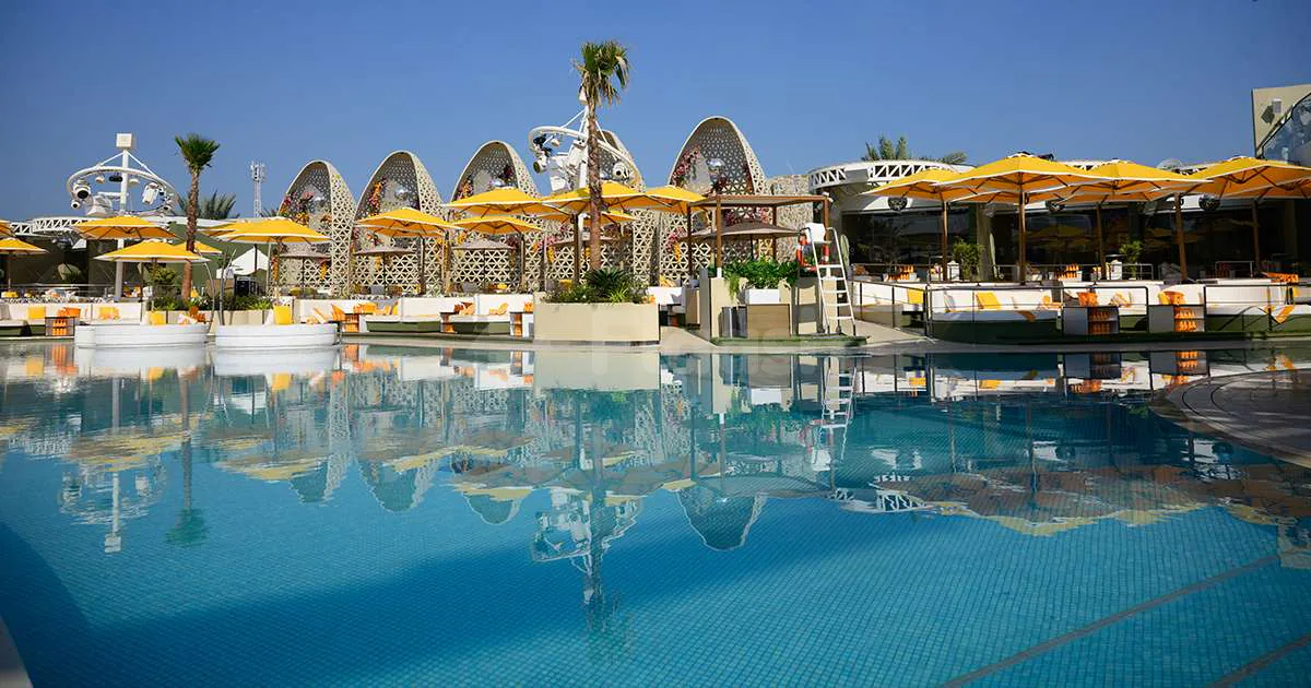 Large swimming pool at O Beach Dubai with yellow umbrellas and curved decorative lattice structures reflected
