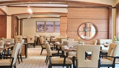 Dining area at Retaj Royale Doha, featuring wooden paneling and a circular mirror above tables set for guests