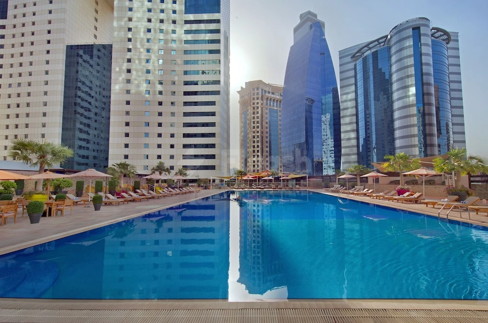 Outdoor swimming pool at Ezdan Hotel & Suites, Doha, reflecting skyscrapers under sunny sky with lounge chairs and umbrellas