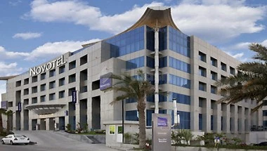 Modern glass facade at Novotel Dammam Business Park, Dammam, with palm trees and blue sky backdrop