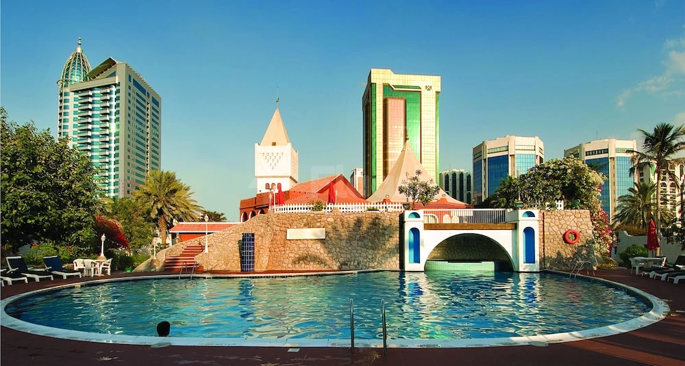 Outdoor pool at Marbella Resort, Sharjah, featuring stone bridge and red-roofed pavilion against city skyline