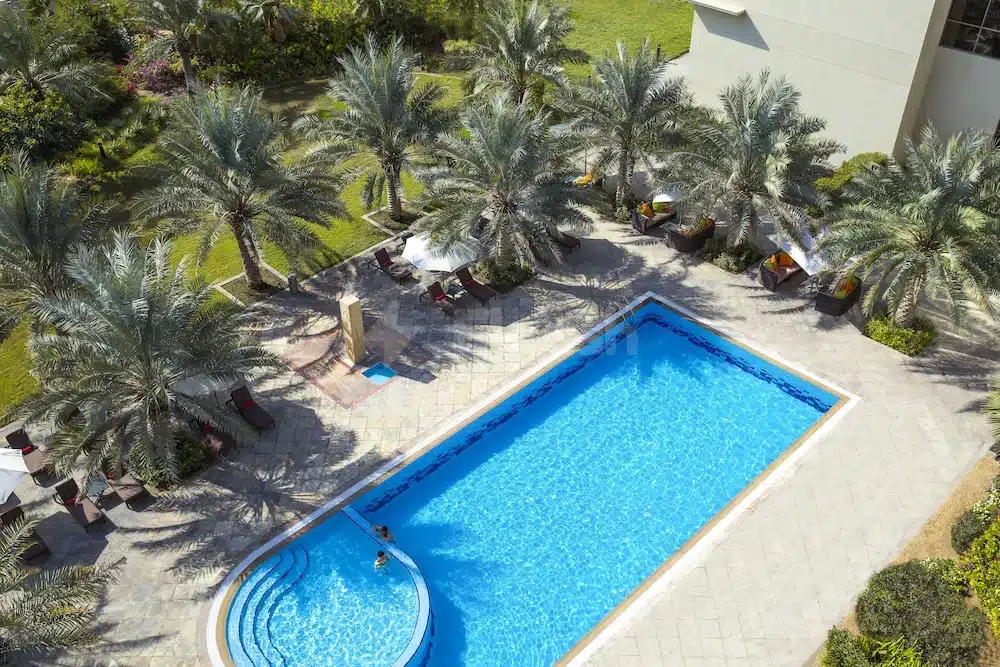 Outdoor swimming pool at Centro Sharjah, Sharjah, surrounded by palm trees and lounge chairs under bright sunlight