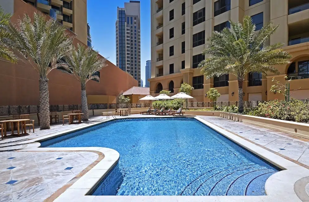 Outdoor pool at Roda Amwaj Suites, Dubai, framed by palm trees and lounge chairs under clear blue sky