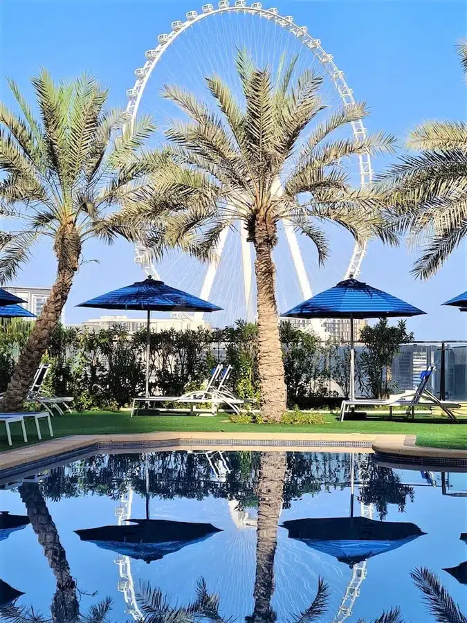 Poolside lounge at Roda Amwaj Suites, Dubai, with blue umbrellas and palm trees reflecting in calm water near
