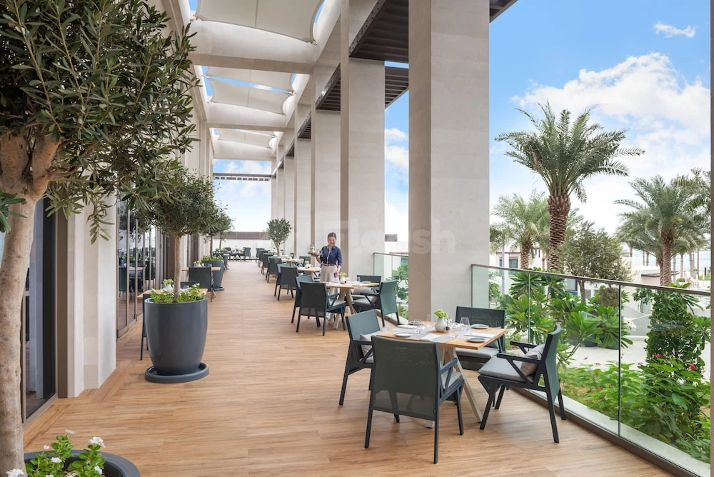 Outdoor dining terrace at Address Beach Resort Fujairah, Fujairah, with olive trees and glass railings framing lush greenery