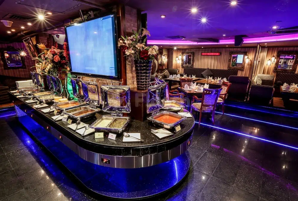 Buffet dining area at Ramee Royal Hotel, Dubai, with blue-lit counter and floral centerpieces under purple ambient lighting