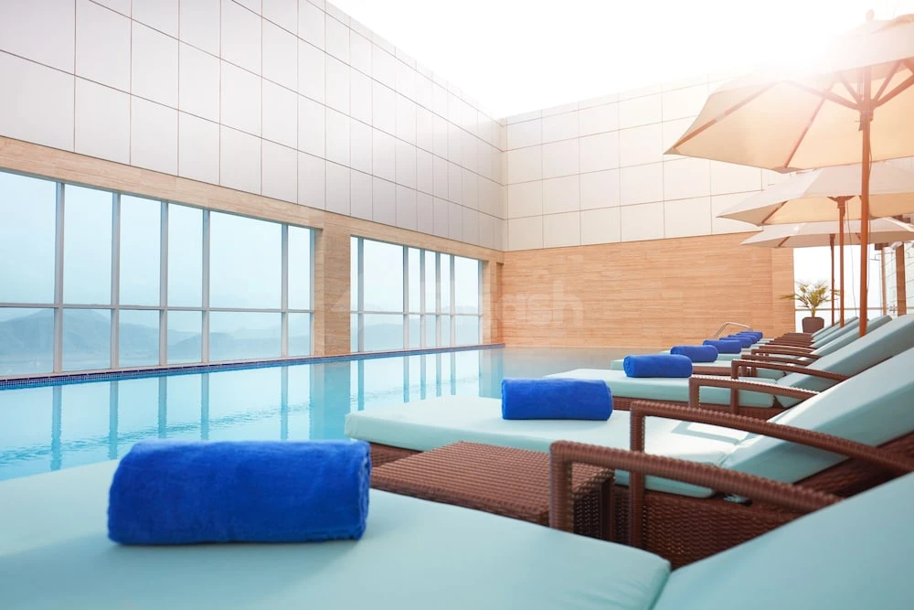 Indoor pool at Royal M Hotel Fujairah, Fujairah, with lounge chairs and blue towels by floor-to-ceiling windows