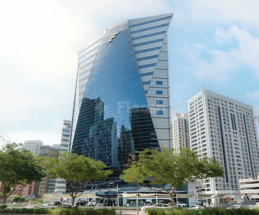 Modern glass skyscraper at Grand Heights Dubai Hotel Apartments, Dubai, with reflective blue facade and surrounding green trees