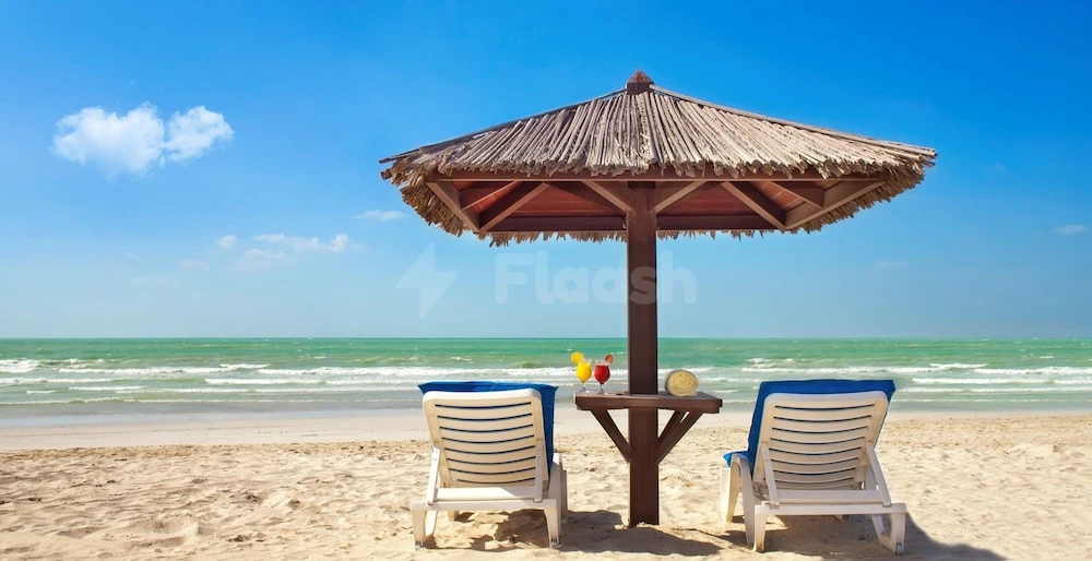 Beach lounge area at Coral Beach Resort,! Sharjah, featuring thatched umbrella and blue cushioned chairs facing turquoise ocean